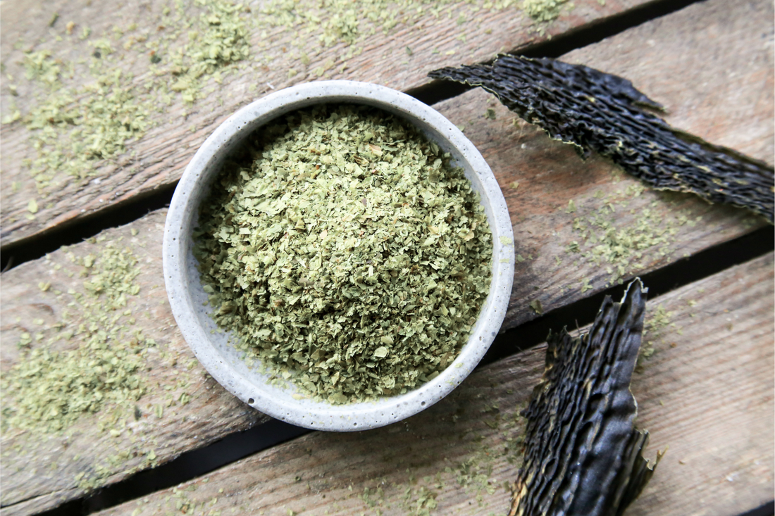 Overhead view of a bowl of finely milled green wakame flakes on a wooden plank surface, with whole dried dark wakame stalks and loose milled flakes scattered nearby